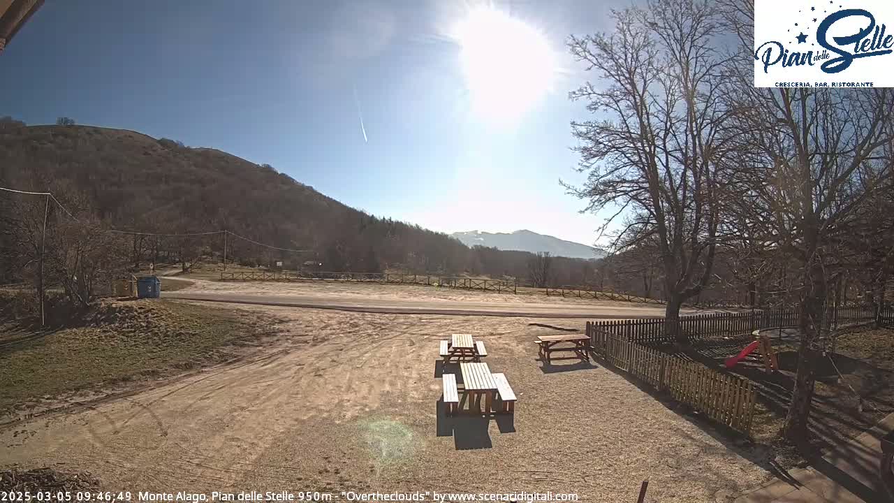 A sunny day with a few clouds reveals a mountain landscape, featuring a gravel area with picnic tables, a small playground, and a road leading to a hill.