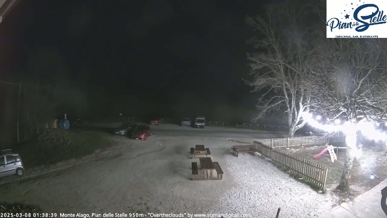 A snow-covered parking lot at night with several cars parked, picnic tables, a small playground, and a tree with string lights.