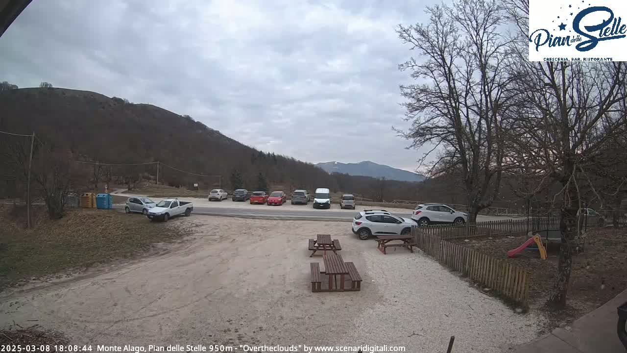 A parking lot with several cars and a few picnic tables is situated in front of a hill under an overcast sky.