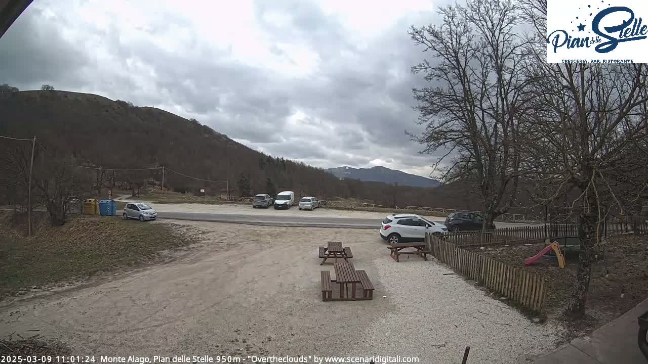 A parking lot with several cars sits near a small playground and picnic tables, overlooking a hillside under an overcast sky.