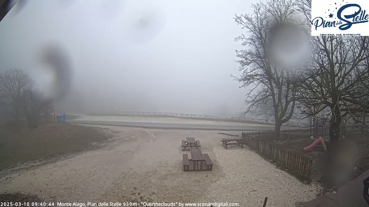A foggy, overcast scene shows a gravel area with picnic tables near a road and a wooden fence, with bare trees in the background.