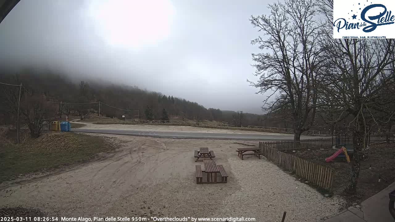 A gravel area with picnic tables and a small playground is situated near a road, overlooking a misty, wooded hillside under an overcast sky.