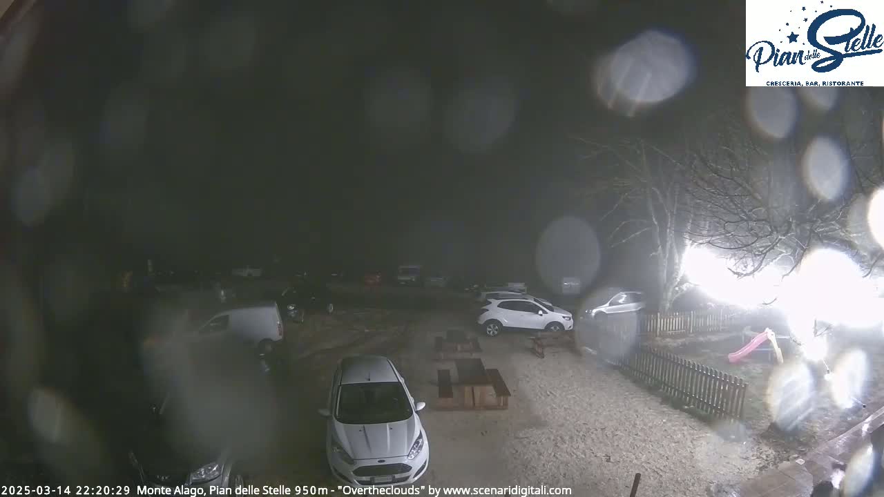 A nighttime, rain-covered parking lot with several cars and a small playground is visible, with some bare trees in the background.