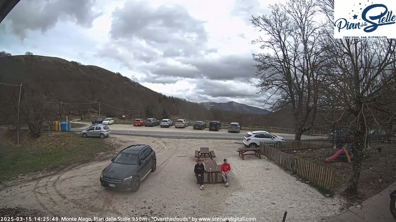 A parking lot with several cars is situated near a small outdoor seating area with two people sitting at a table, all under an overcast sky in a mountainous area.