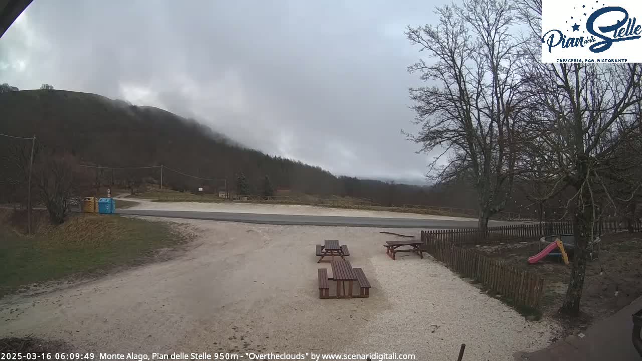 A parking area with picnic tables sits near a road, overlooking a mountain shrouded in overcast weather.