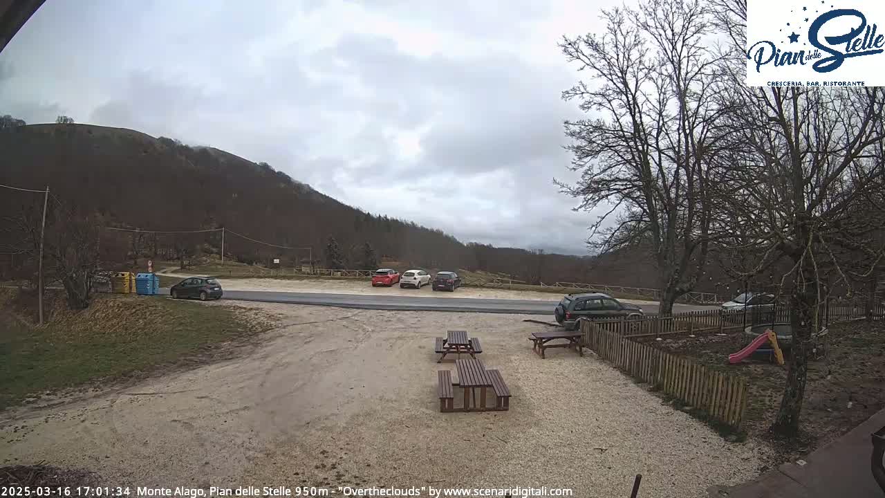 A parking lot with several cars sits before a small picnic area next to a wooden fence, all overlooking a mountain under an overcast sky.