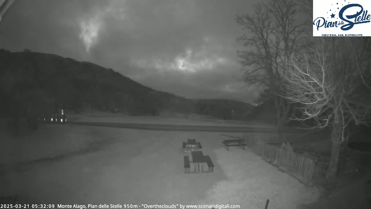 A nighttime, overcast scene shows several empty picnic tables in a snow-covered area near bare trees and a dark hill in the background.