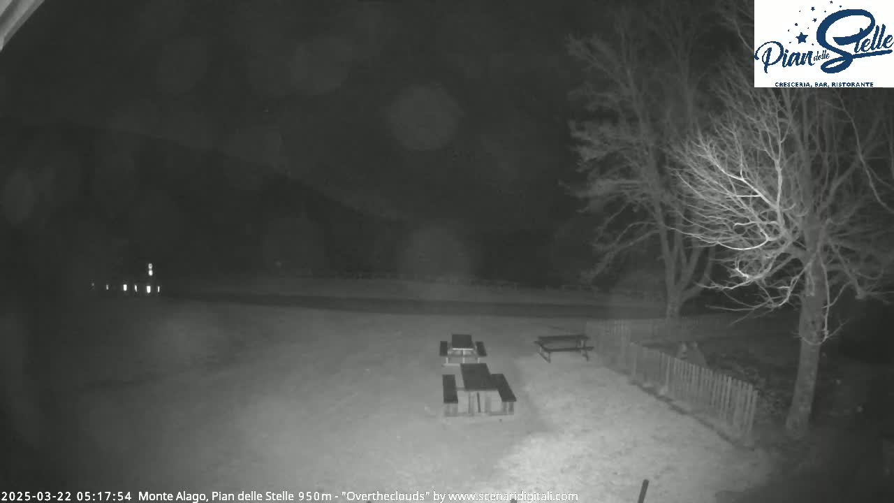 A nighttime, snow-covered area with several picnic tables and bare trees under an overcast sky.