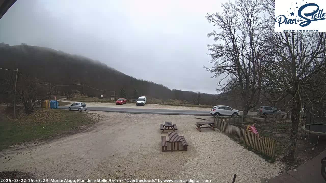 A parking lot with several cars sits below a misty, overcast mountainside, with picnic tables and a small playground visible in the foreground.
