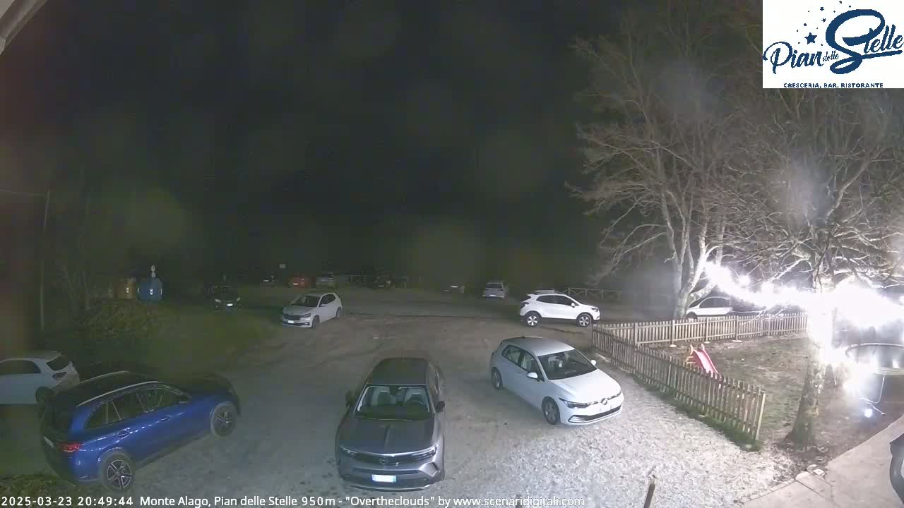 A nighttime parking lot with several cars, illuminated by lights strung along a nearby fence, under an overcast sky.