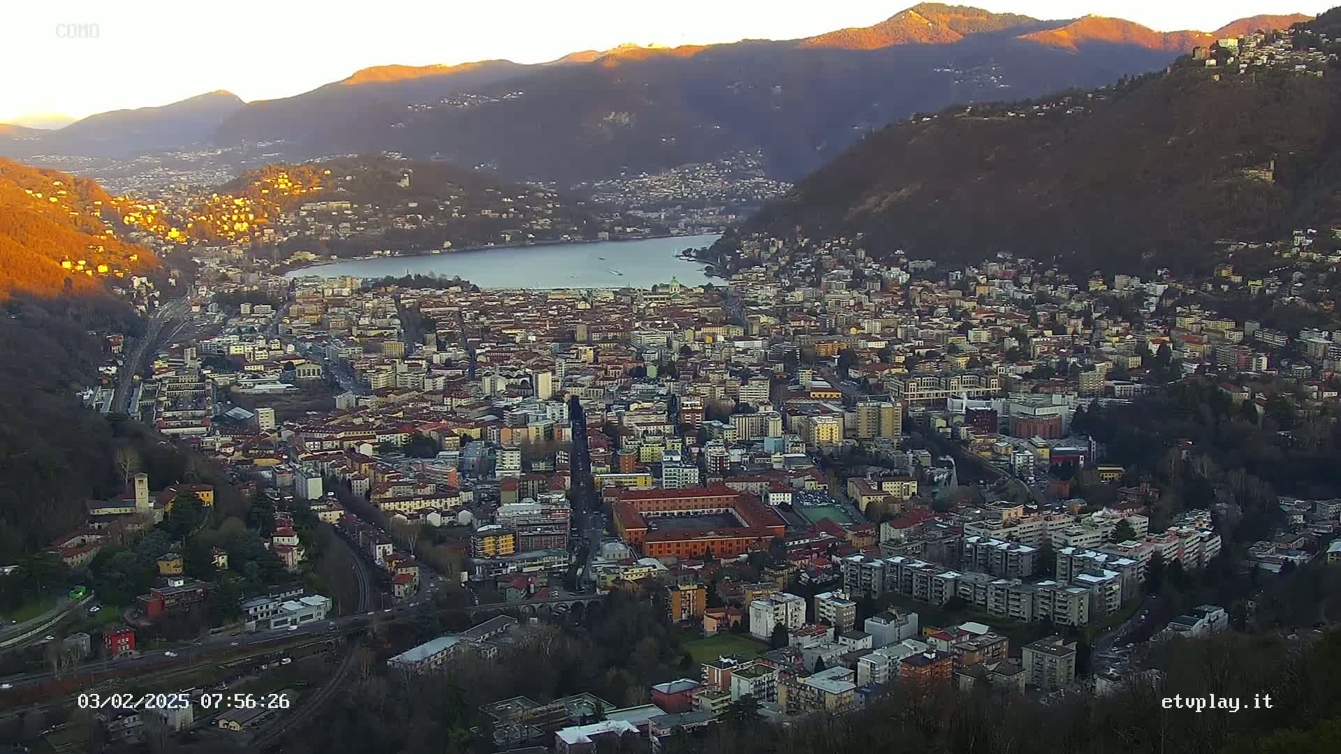 A sprawling city nestled between mountains and a lake at sunrise, the sun illuminating the mountaintops and the city's buildings.
