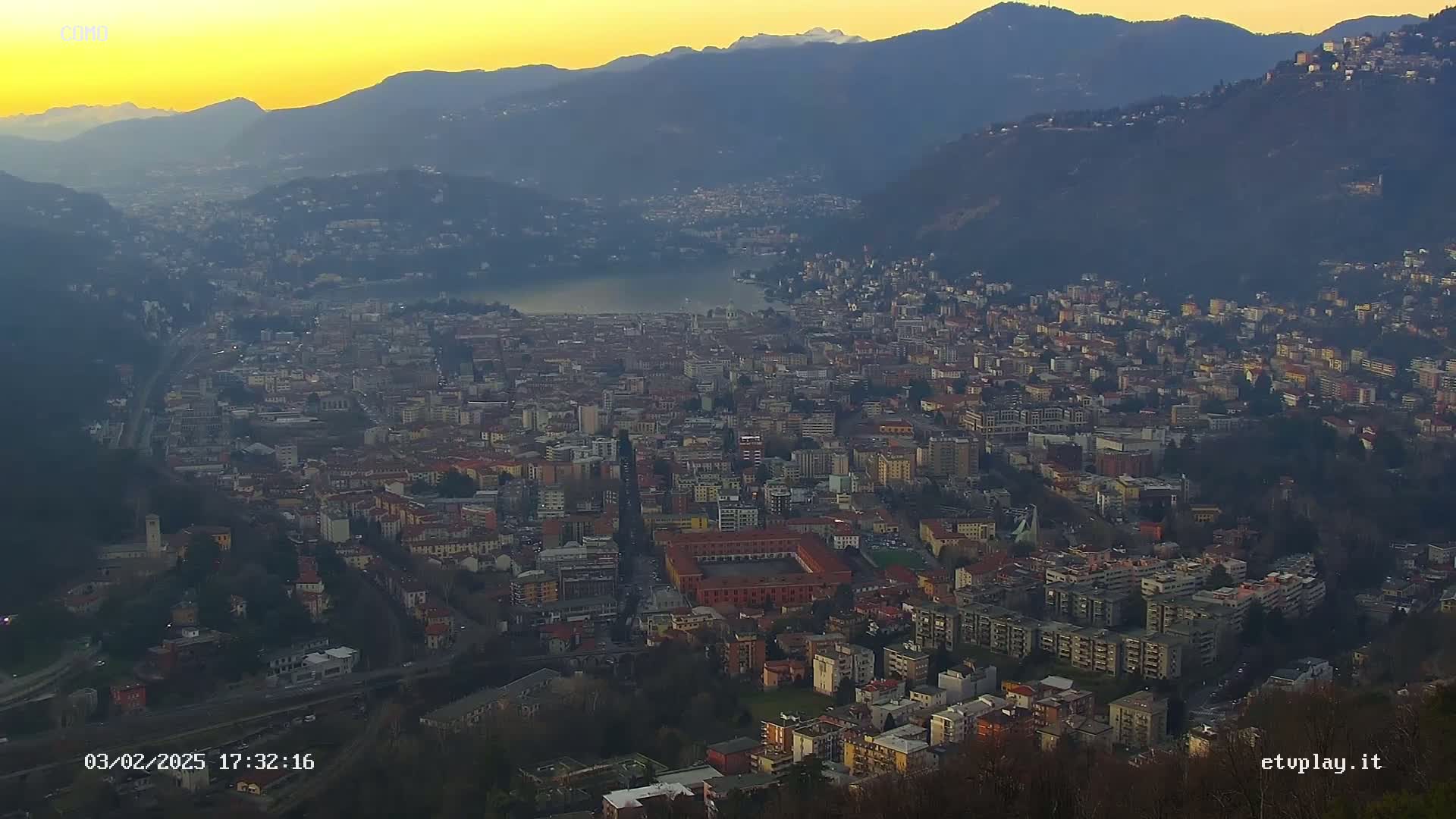 A sprawling city nestled in a valley at sunset, with mountains in the background and a lake partially visible.