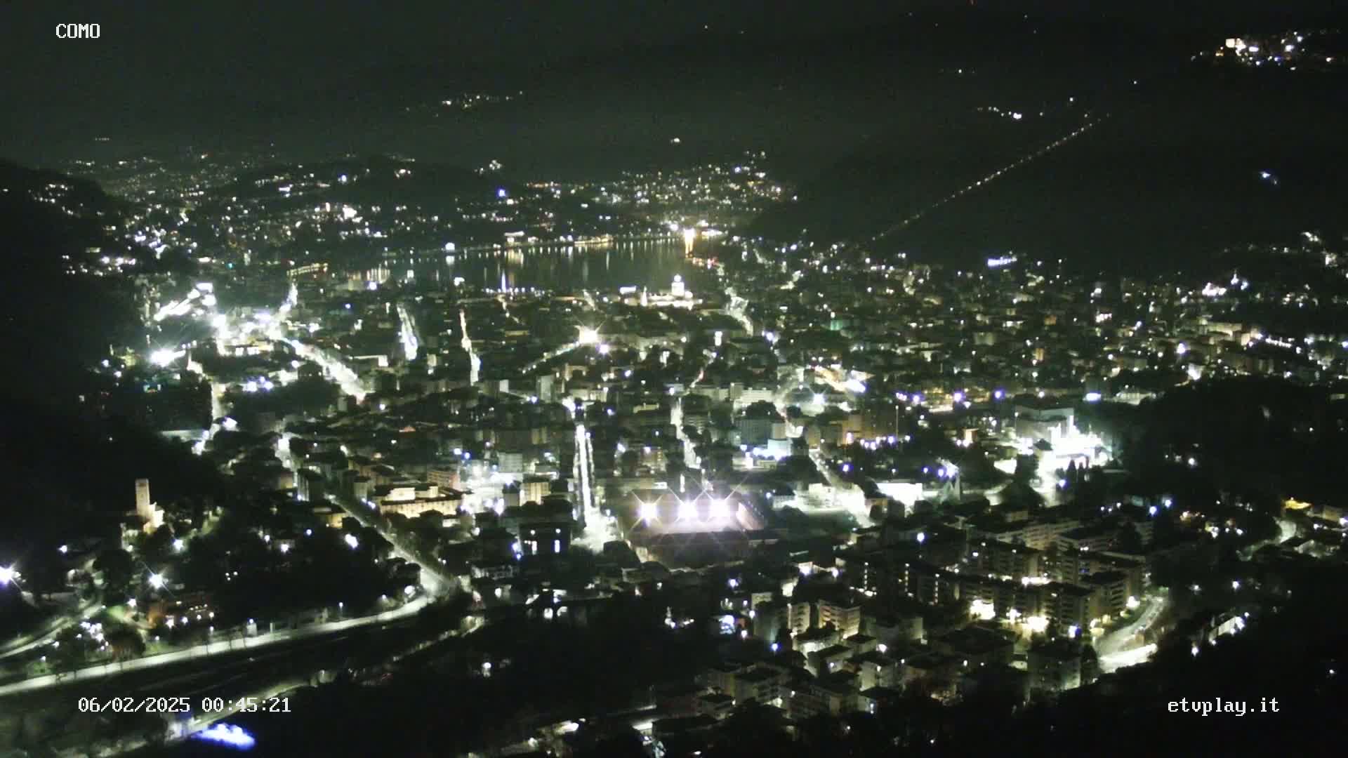 A nighttime, aerial view of a city illuminated by streetlights, nestled beside a dark body of water under a clear sky.