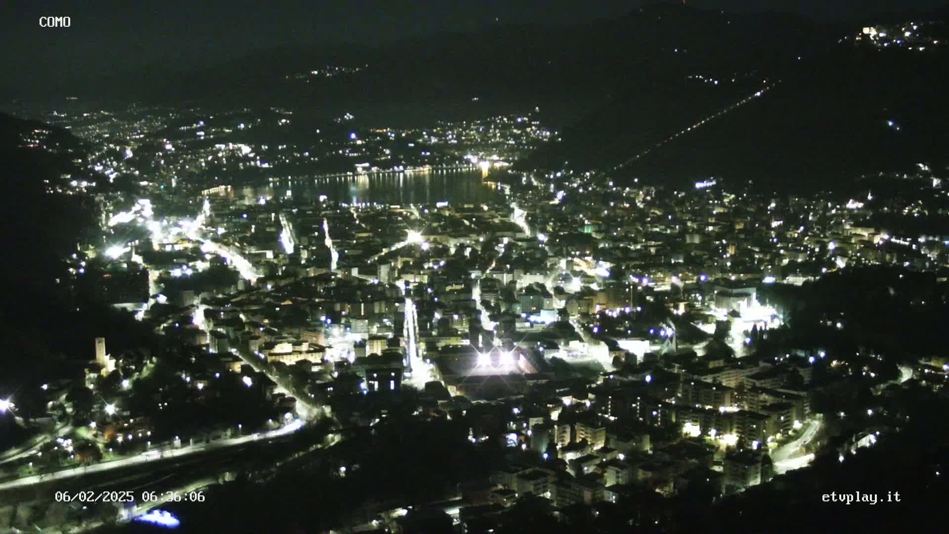 A nighttime aerial view of a city illuminated by streetlights, situated beside a dark body of water under a clear sky.