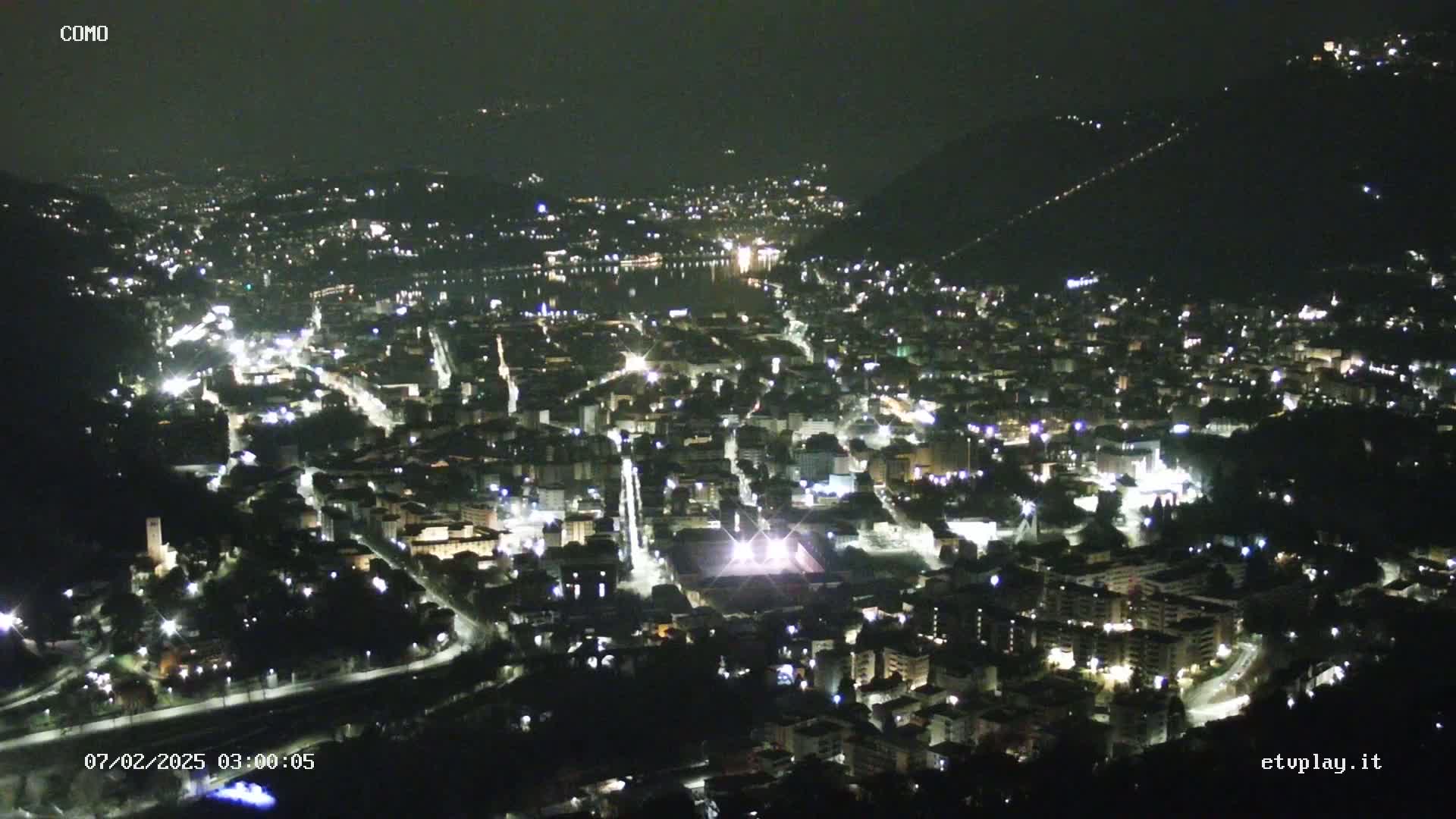 A nighttime aerial view of a city nestled in a valley, with its lights illuminating the streets and buildings under a clear sky.