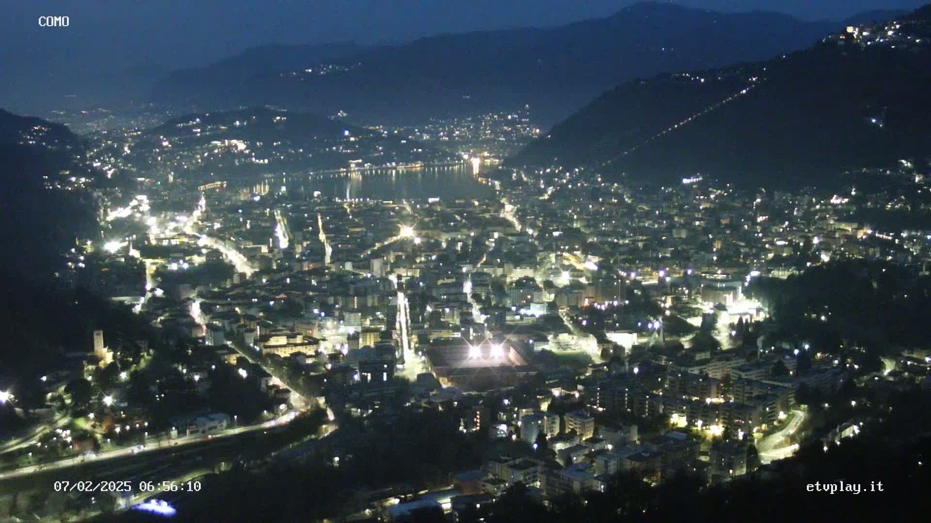 A nighttime, high-angle view of a city illuminated by streetlights, nestled between hills and a body of water.