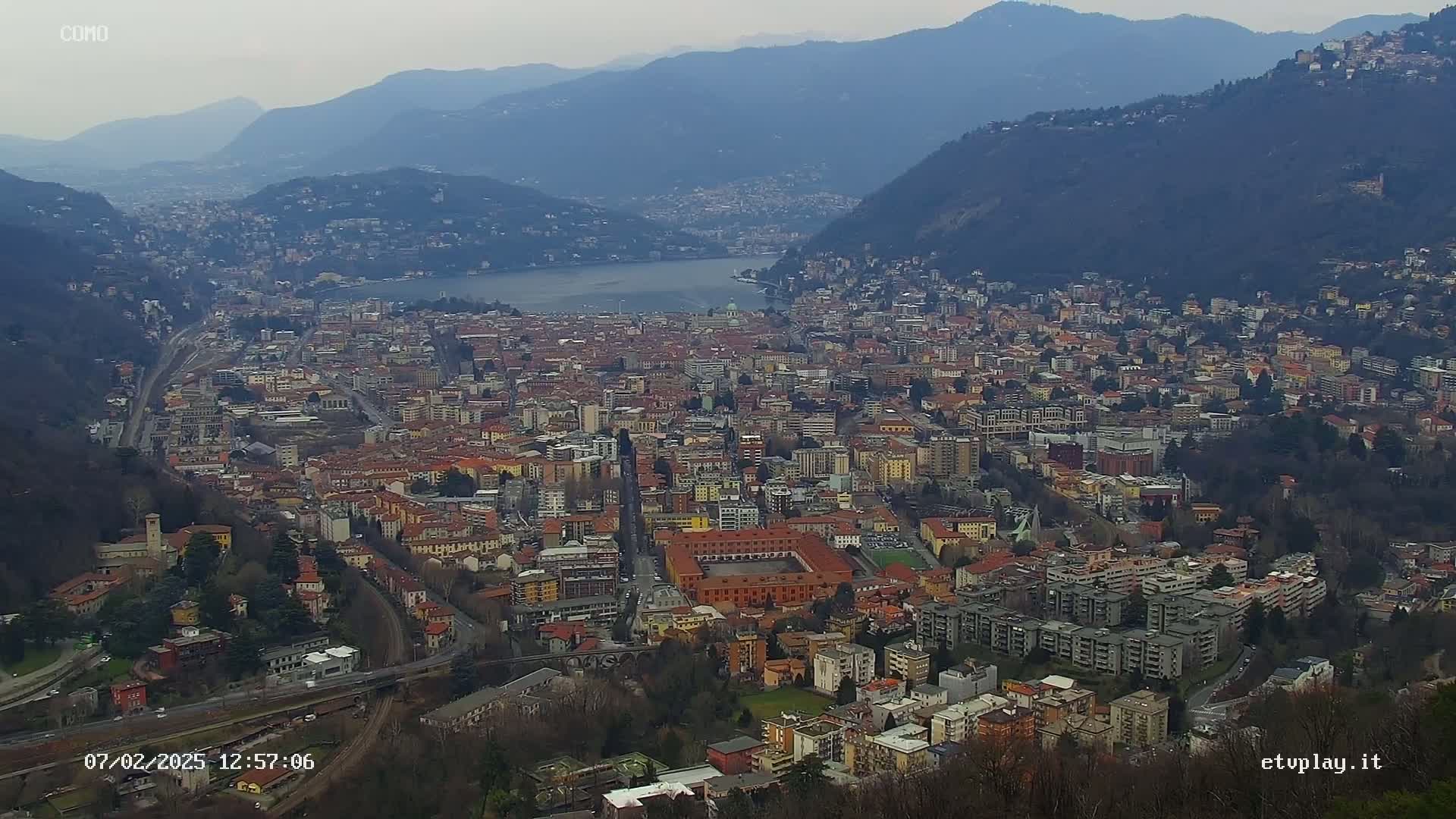 A sprawling city nestled between mountains and a lake on an overcast day.