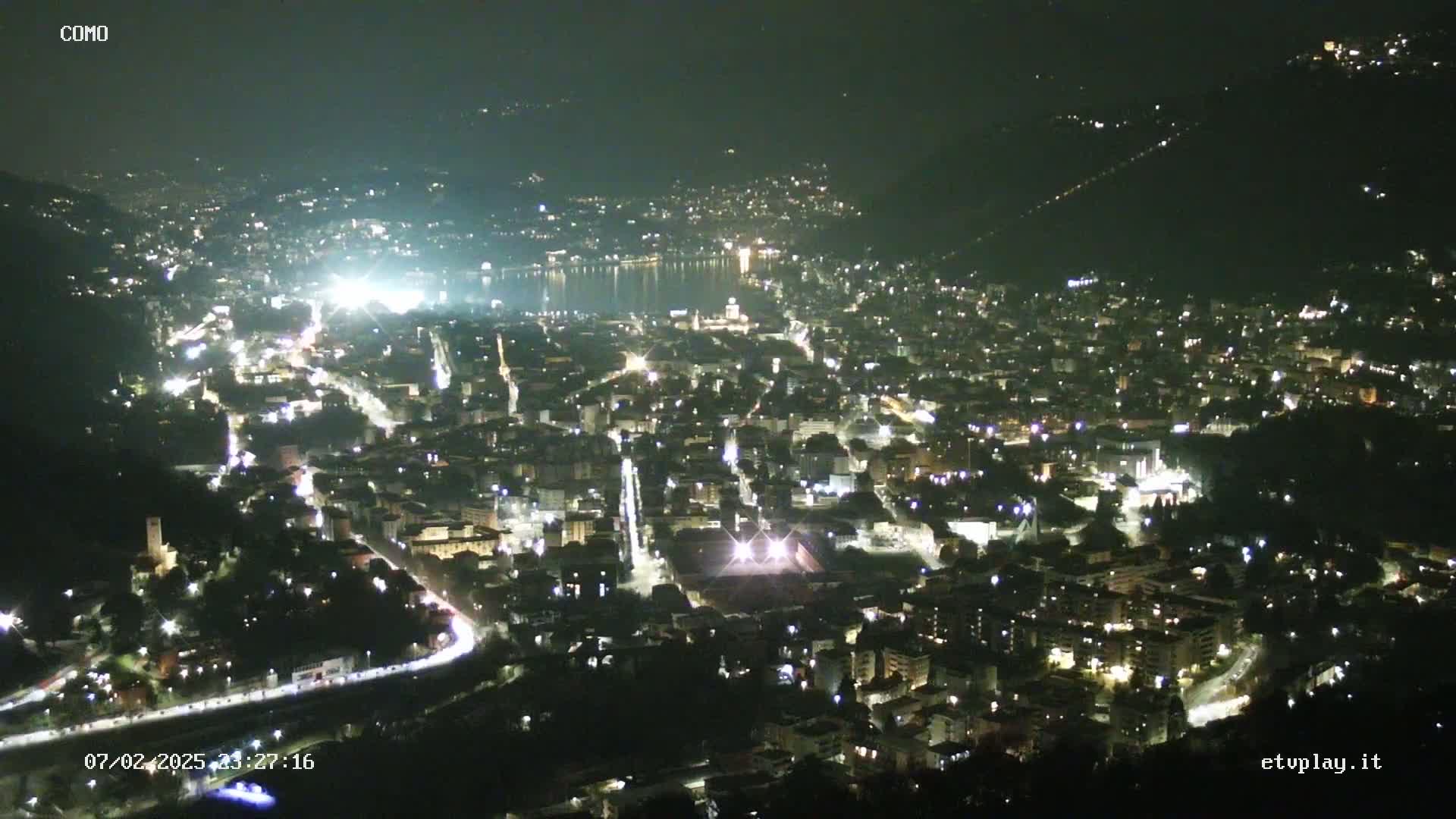 A nighttime, aerial view shows a city illuminated by lights, nestled beside a body of water, under a clear, dark sky.