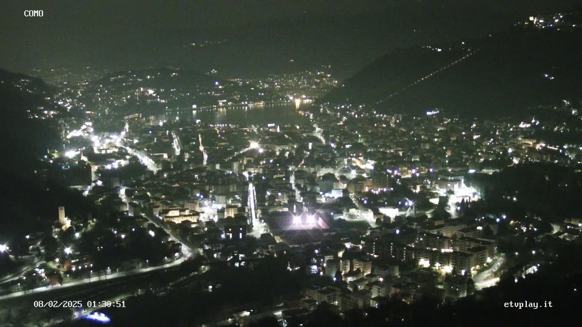 A nighttime aerial view of a city situated near a body of water, with the city's lights illuminating the landscape under a clear, dark sky.
