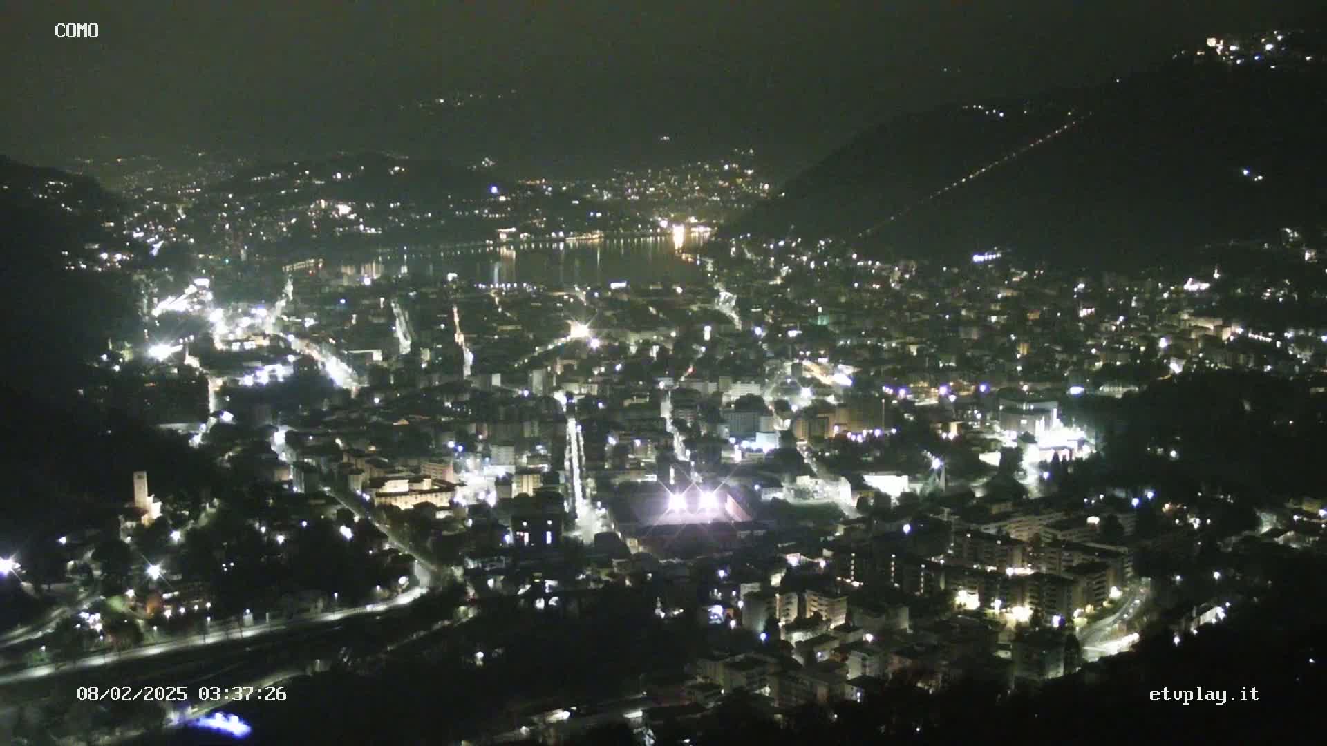 A nighttime aerial view of a city illuminated by lights, situated beside a dark body of water under a clear, dark sky.