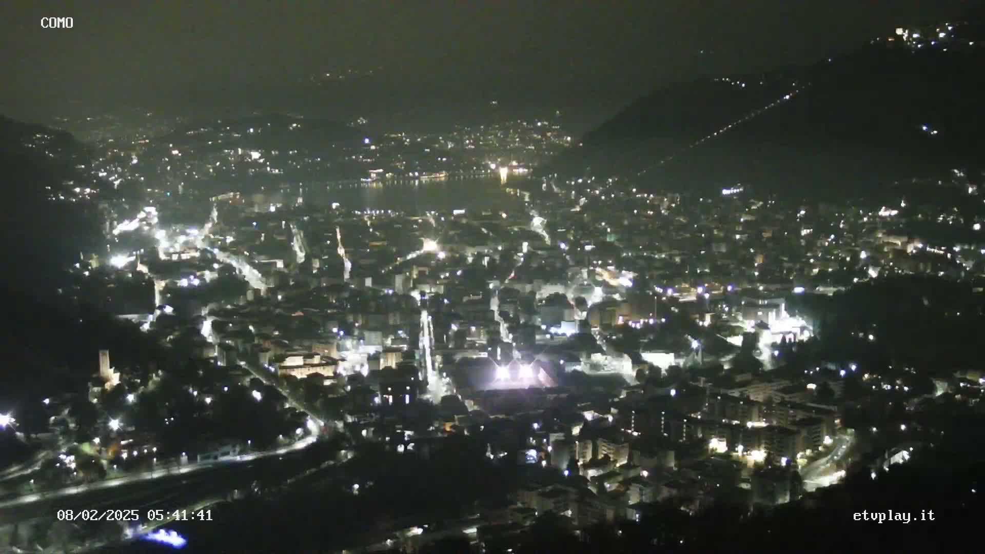 A nighttime, aerial view of a city nestled between hills and a body of water, illuminated by streetlights and building lights.
