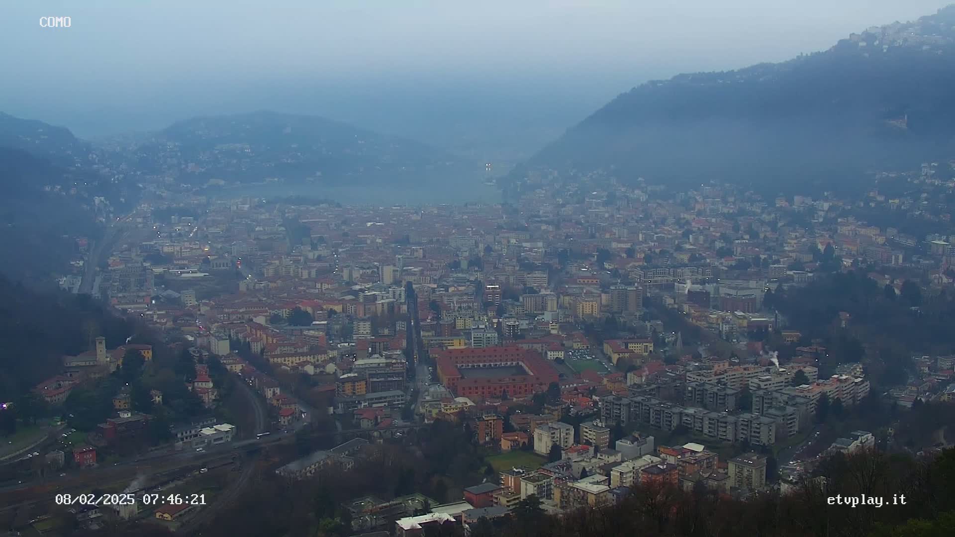 A hazy, low-visibility view from a high vantage point shows a sprawling city nestled in a valley between hills.