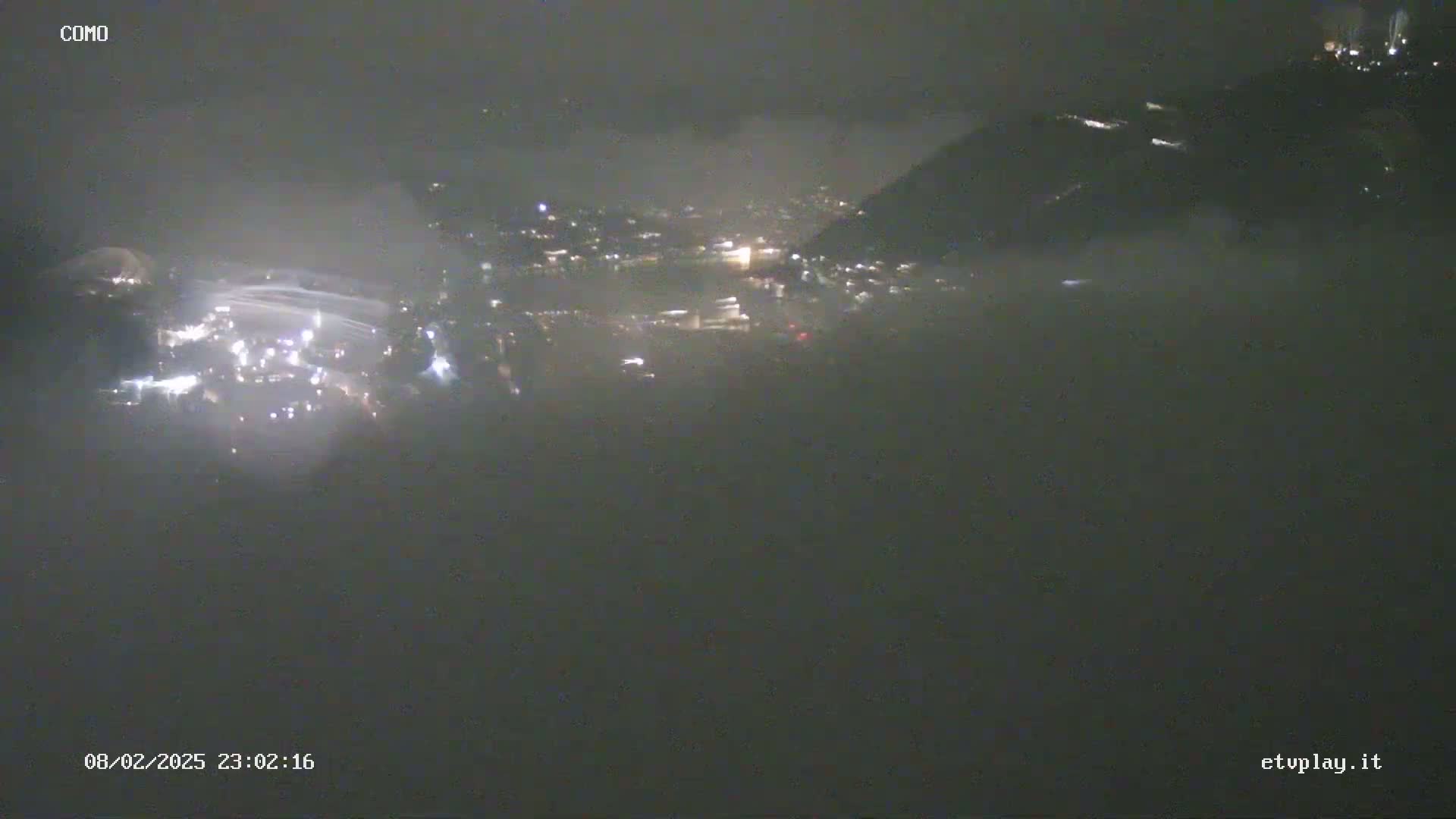 A nighttime, foggy view shows a city illuminated by scattered lights below a dark, hilly landscape.