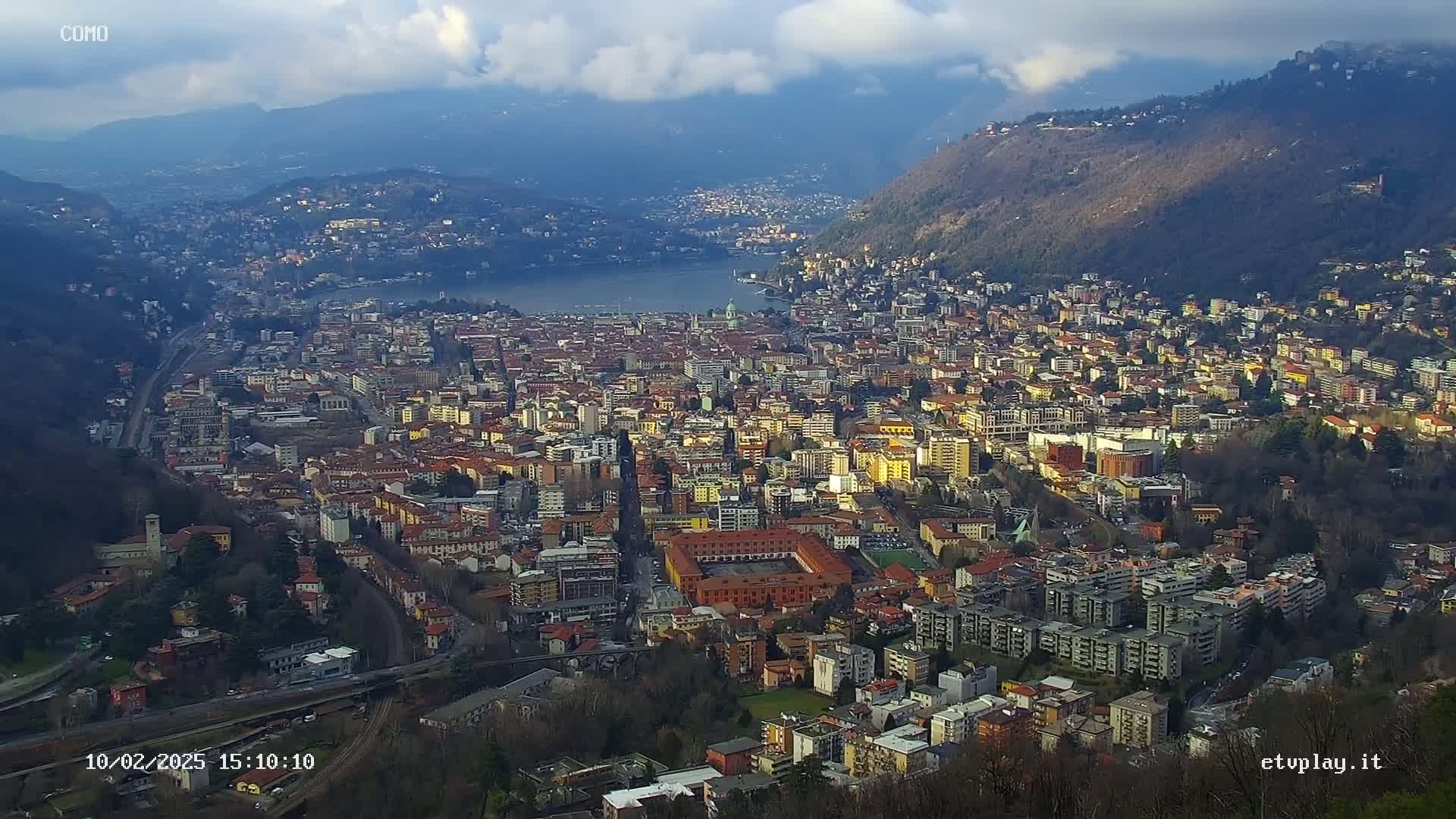 An expansive view of a city nestled beside a lake, surrounded by mountains under a partly cloudy sky.