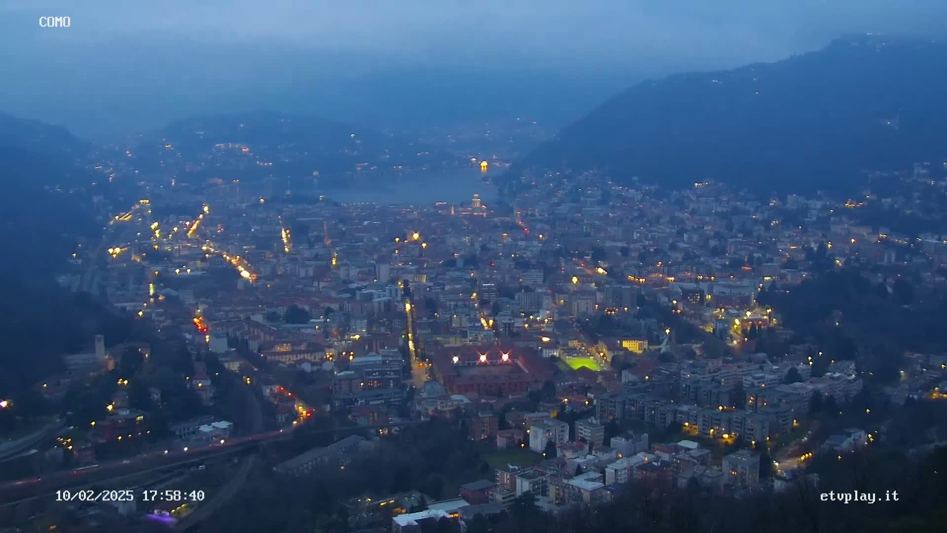 A nighttime, aerial view of a city nestled in a valley, illuminated by streetlights and with a body of water visible in the distance under a dusky sky.