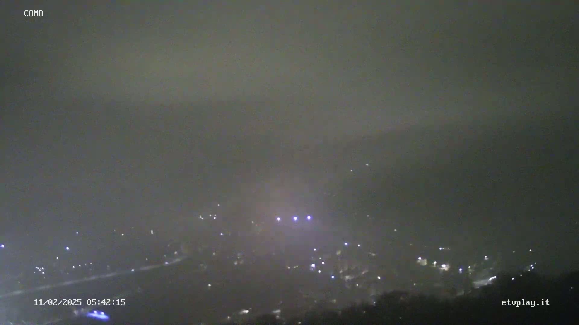 A hazy, nighttime view shows a scattering of faint lights across a landscape, possibly a town or city, under a dark, overcast sky.