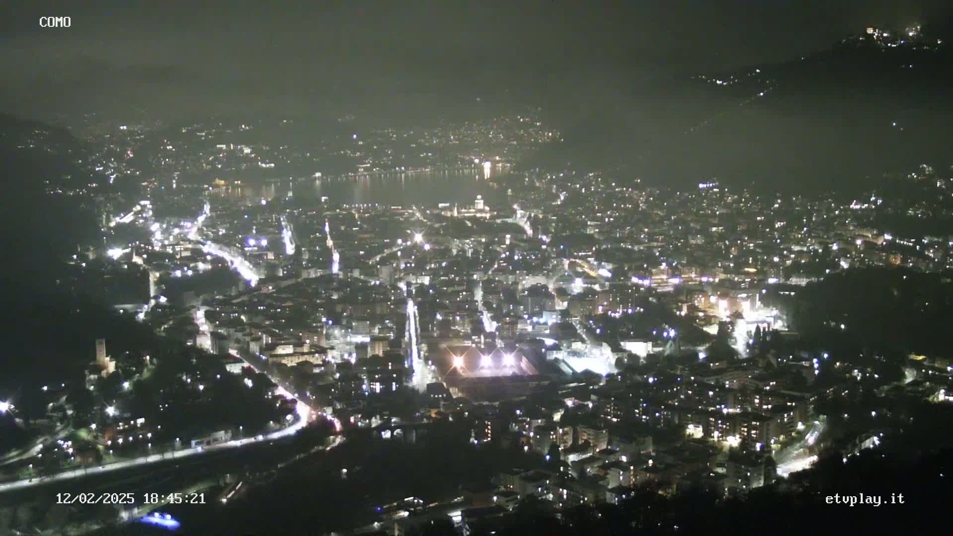 A nighttime aerial view of a city illuminated by lights, situated near a body of water under a dark, clear sky.