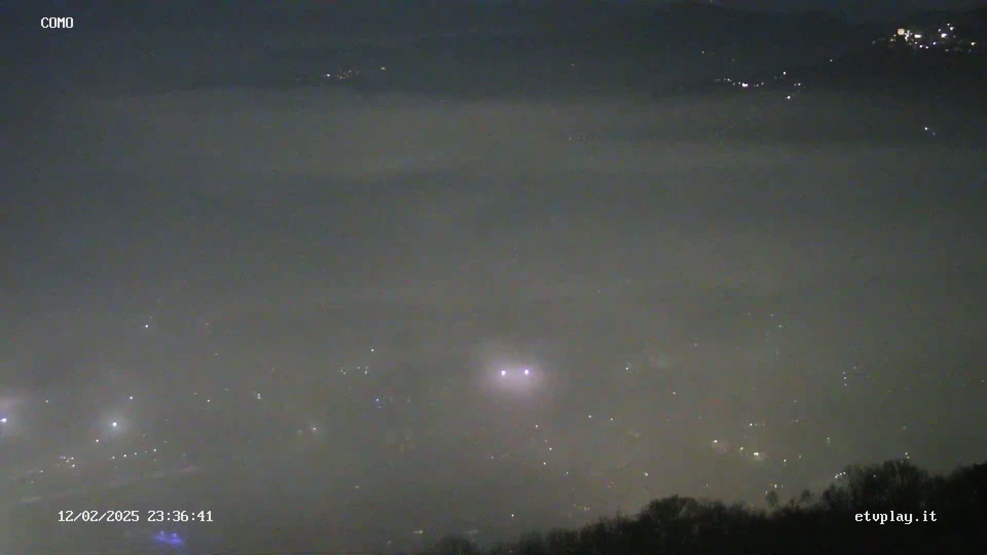 A nighttime, foggy landscape shows distant lights scattered across a valley below a hill covered in dark trees.