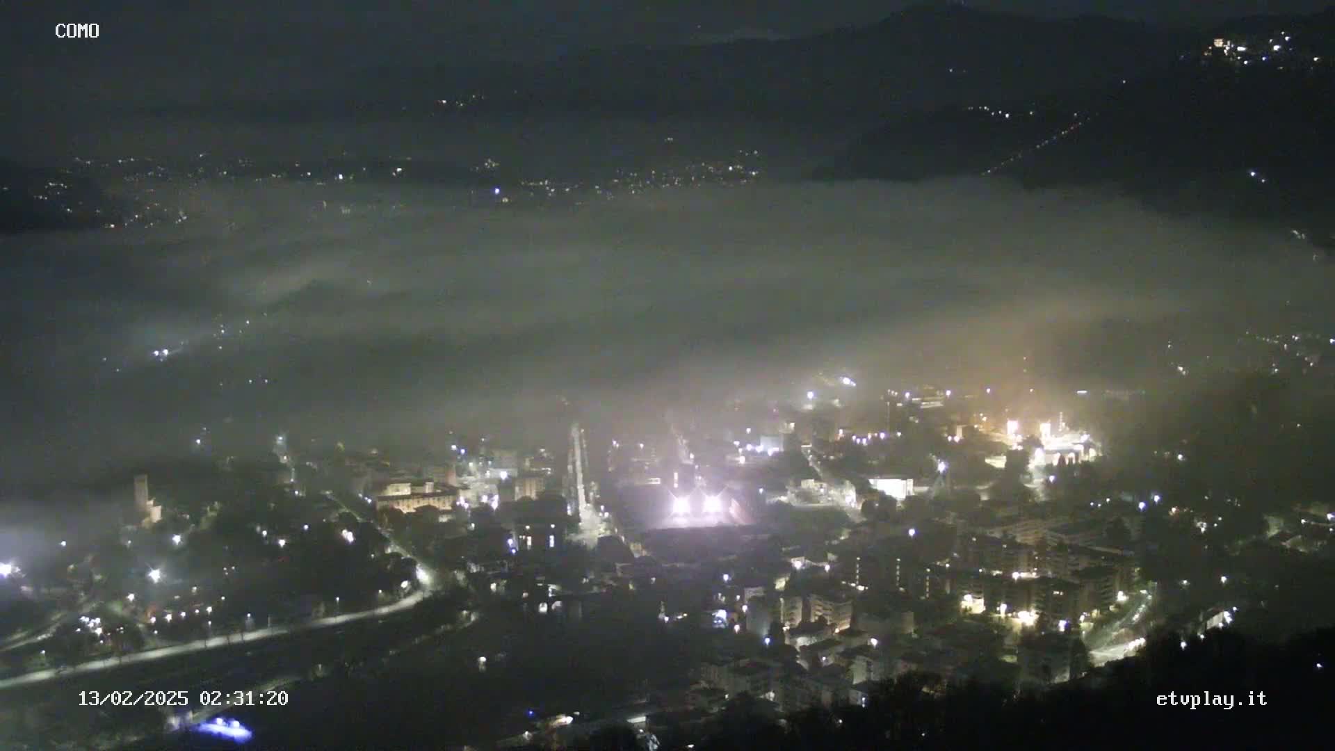 A nighttime, foggy view of a city nestled in a valley, with lights illuminating the buildings and streets below.