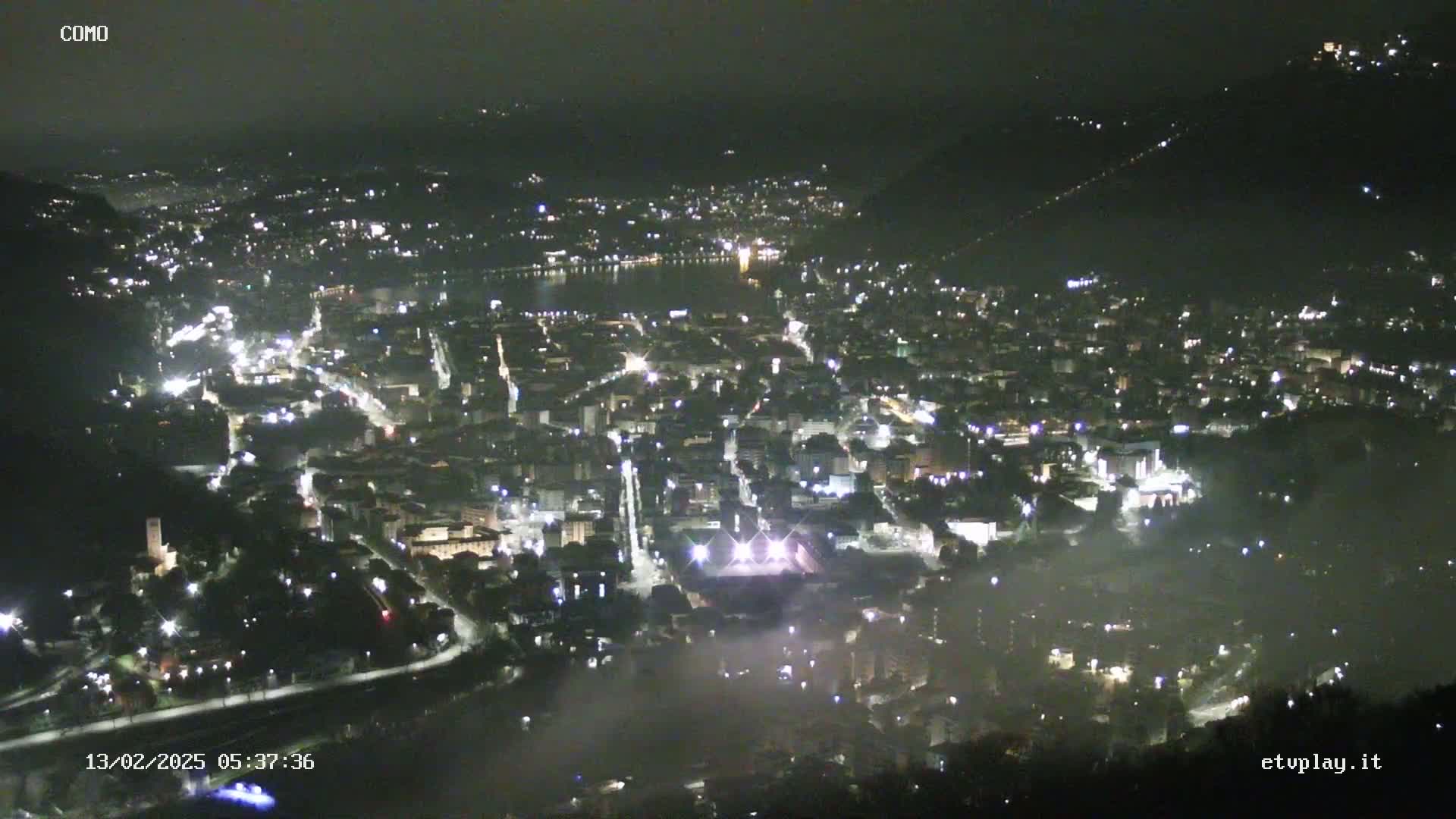 A nighttime, hazy aerial view of a city illuminated by streetlights and building lights, with a body of water visible in the distance.