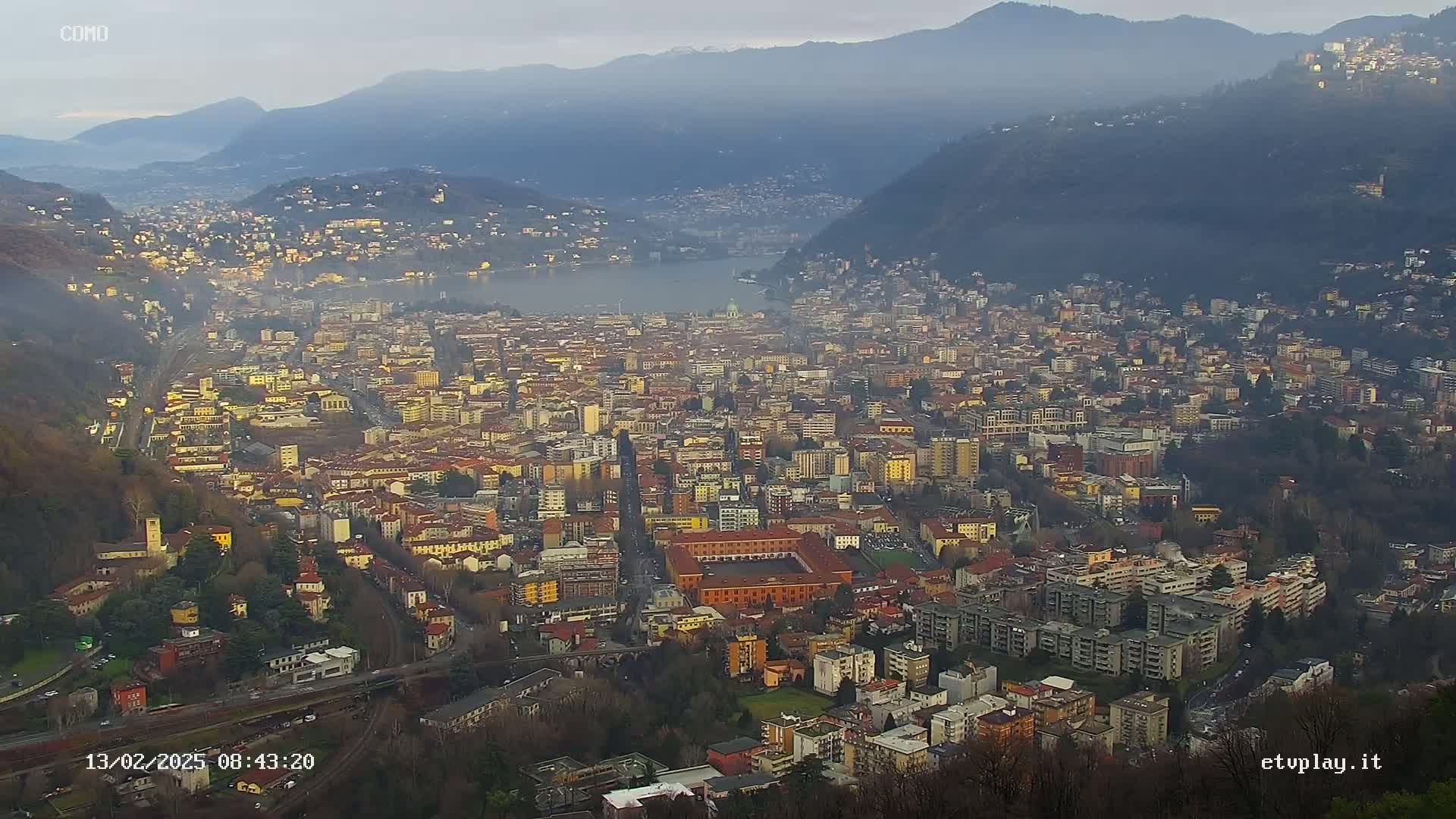 A hazy, panoramic view of a large city nestled in a valley between mountains, overlooking a lake.