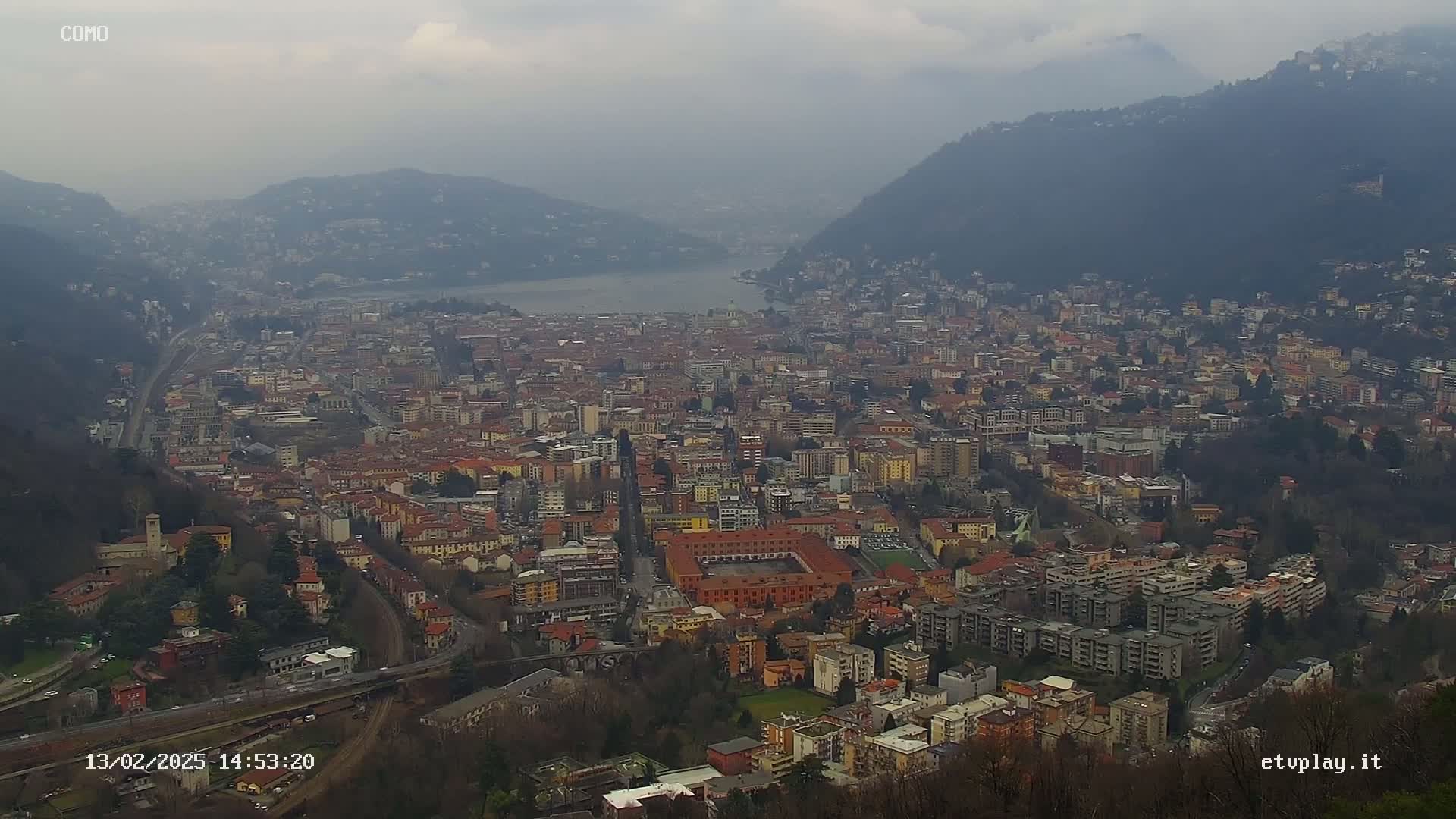 A sprawling city nestled between mountains and a lake under a cloudy sky.