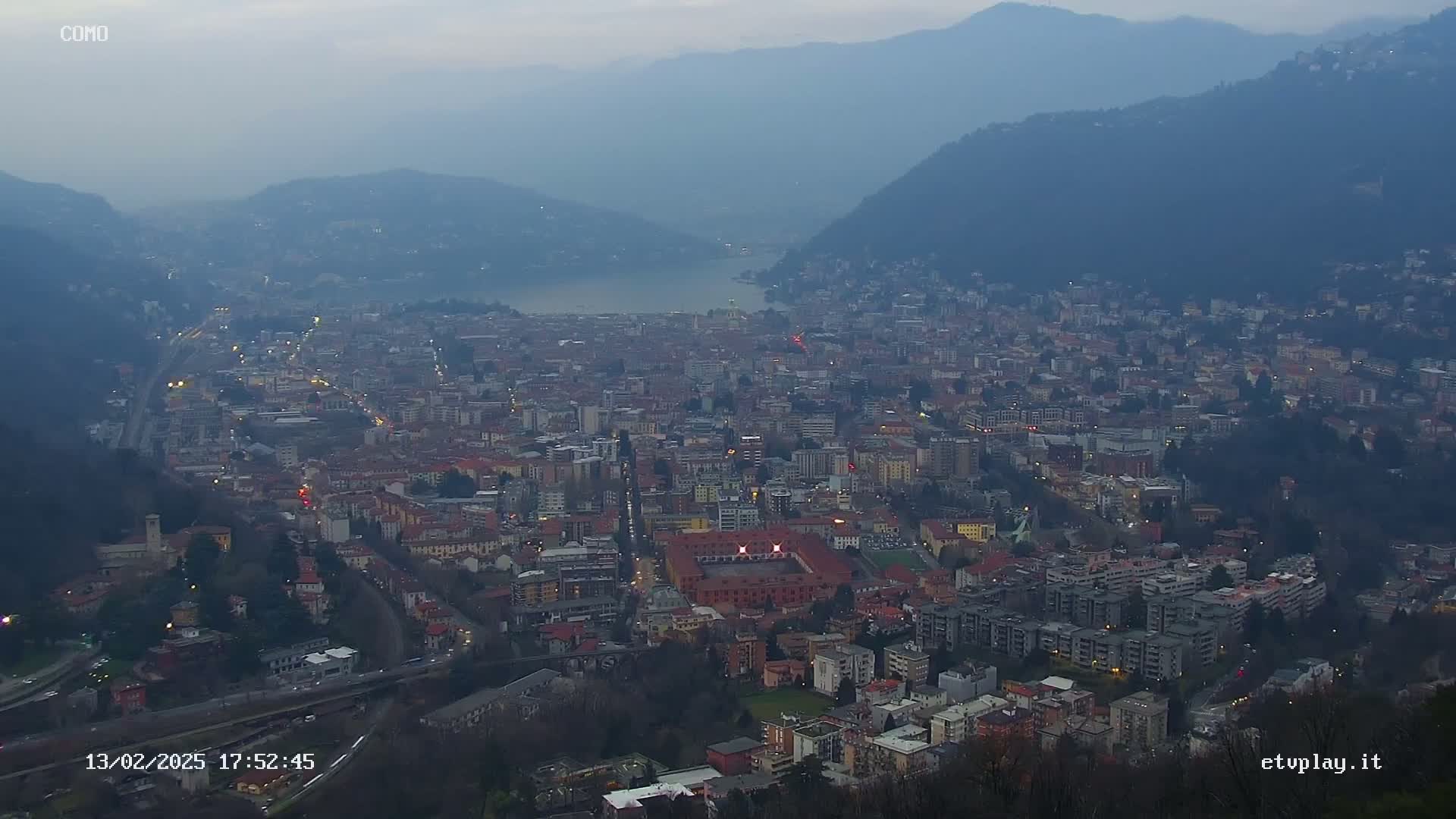 A sprawling city nestled between mountains and a lake at dusk under a hazy sky.