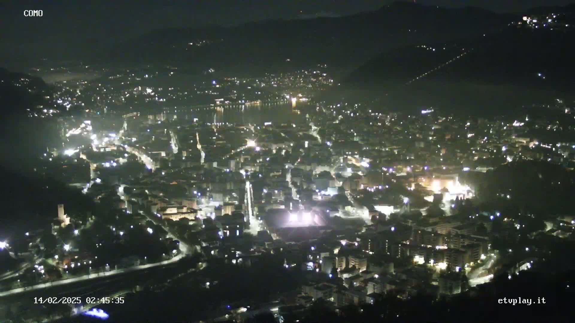 A nighttime aerial view of a city nestled beside a body of water, illuminated by lights, under a clear, dark sky.