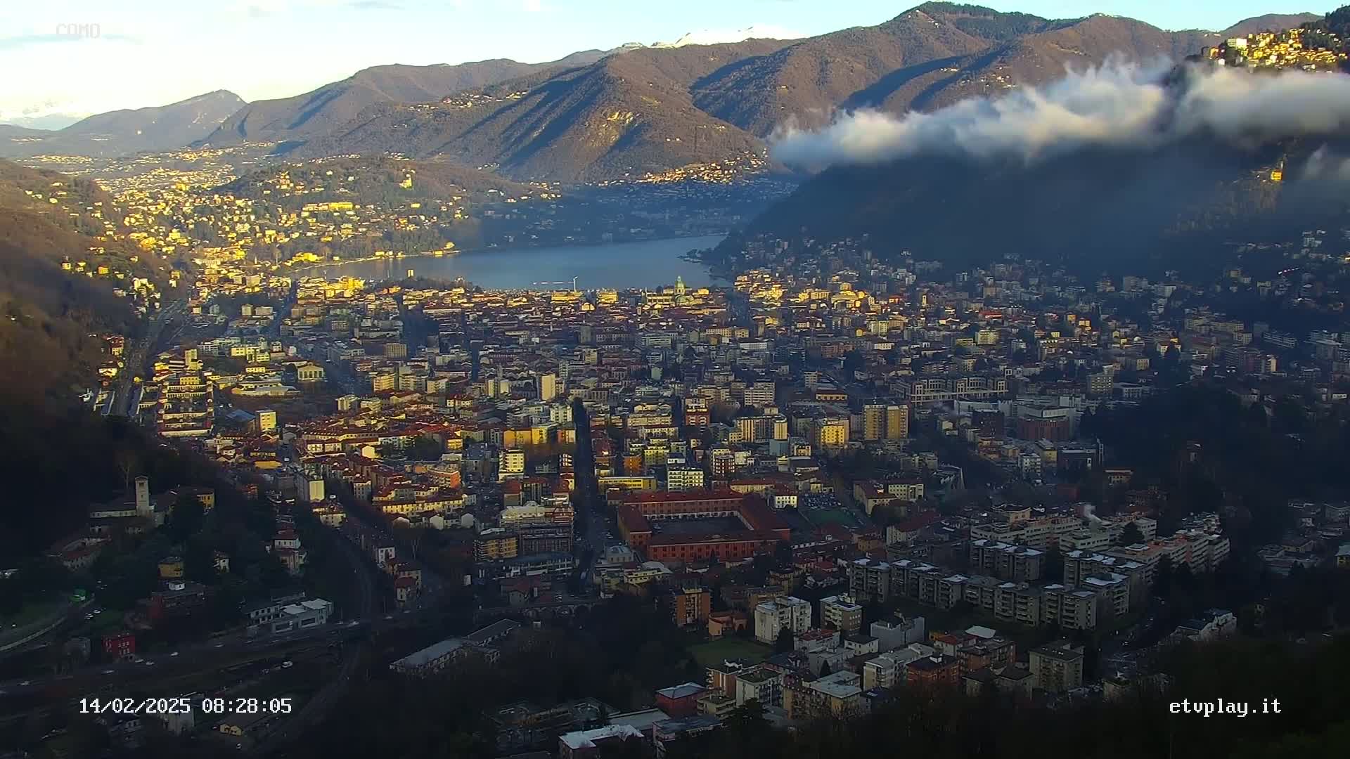 A sprawling city nestled between mountains and a lake, partially shrouded in fog under a mostly clear sky.