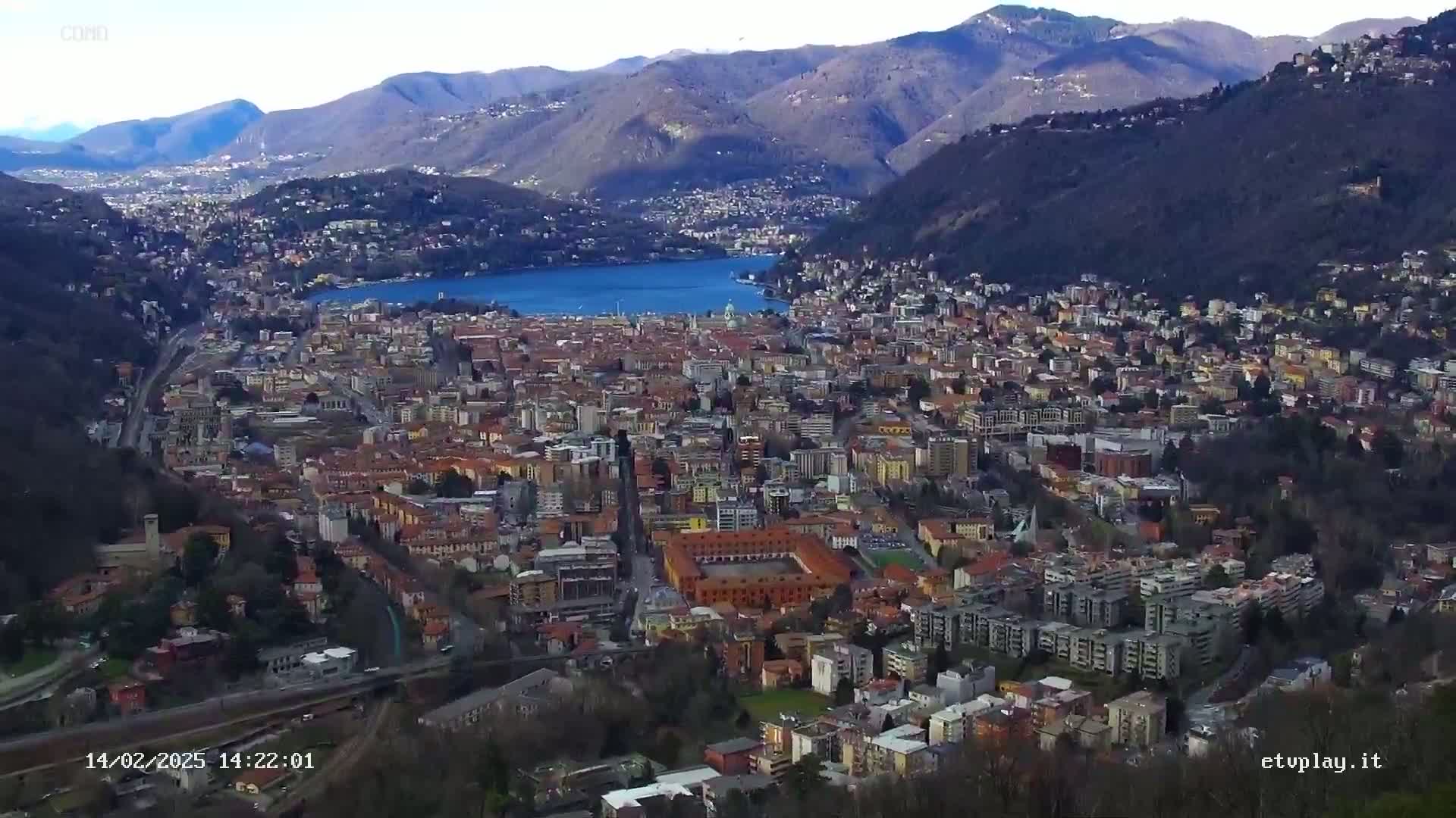 A sprawling city nestled in a valley beside a large lake, surrounded by mountains under a clear, sunny sky.
