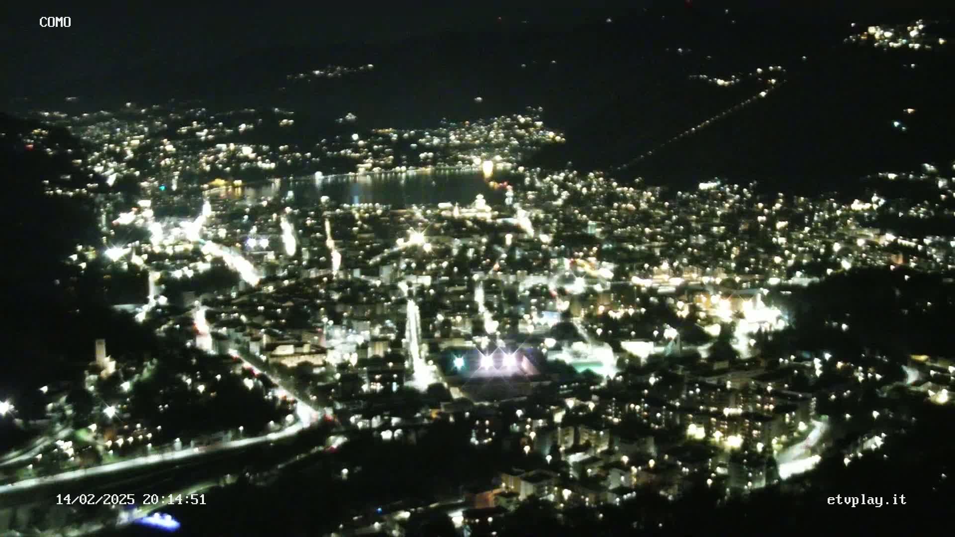 A nighttime aerial view of a city illuminated by numerous lights, situated beside a dark body of water under a clear sky.
