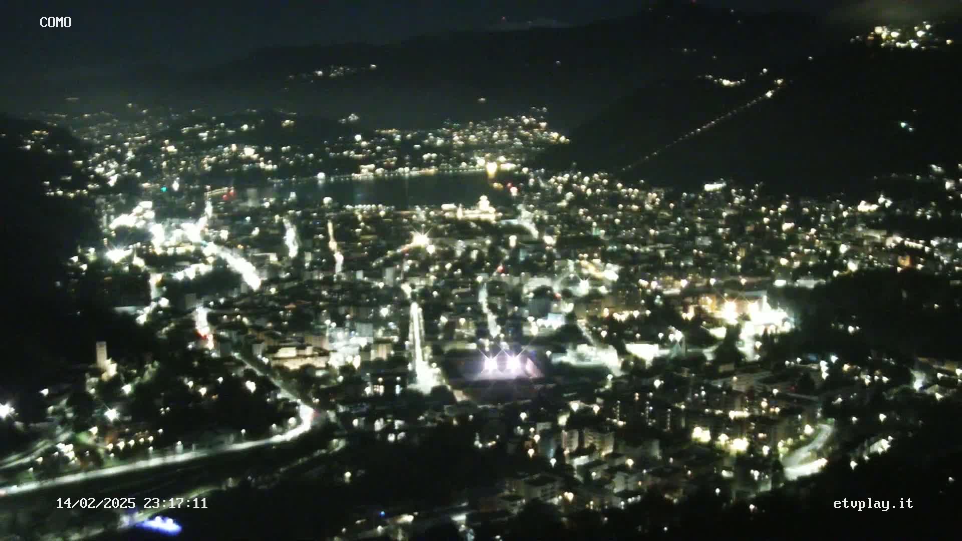 A nighttime, aerial view of a city illuminated by many lights, situated on a hillside overlooking a dark body of water.