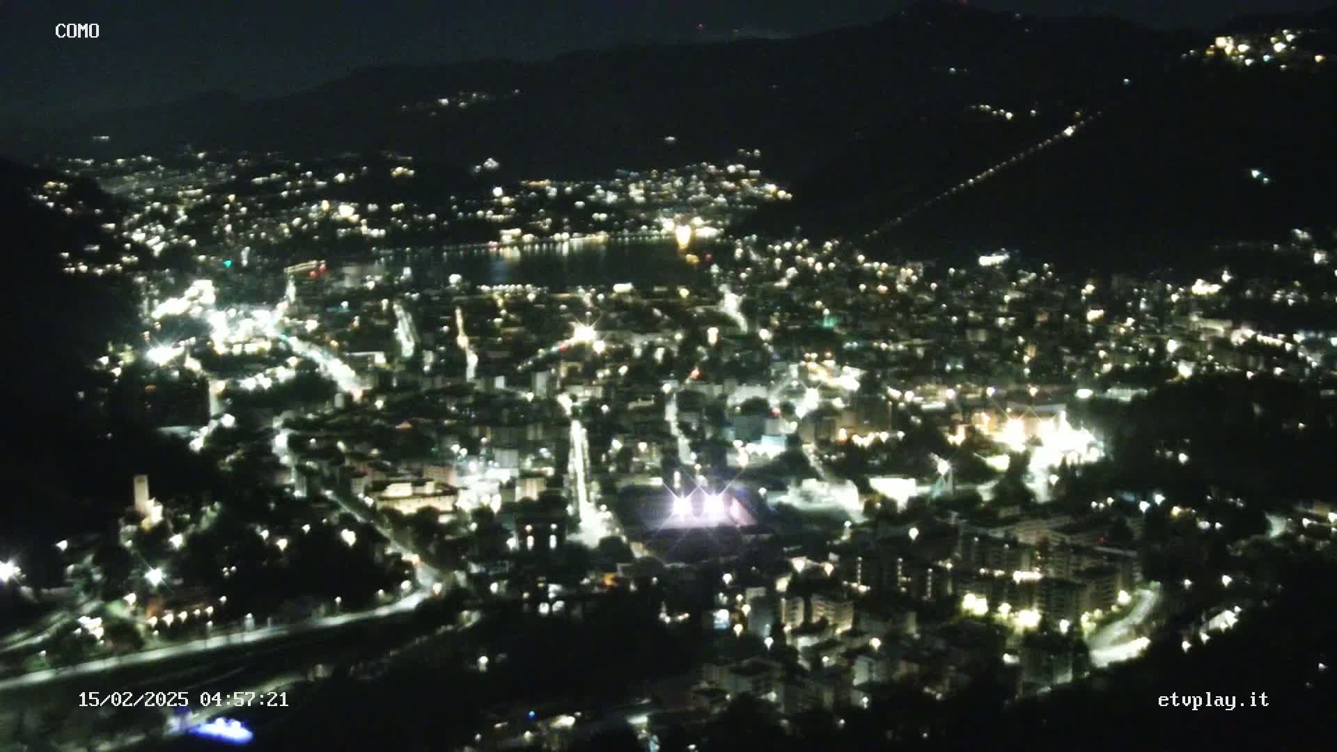 A nighttime, high-angle view shows a city illuminated by numerous lights, situated on a hillside next to a dark body of water.