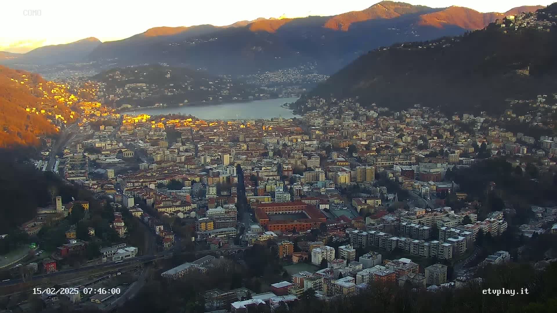 A sprawling city nestled in a valley between mountains at sunrise, with a lake visible in the distance.