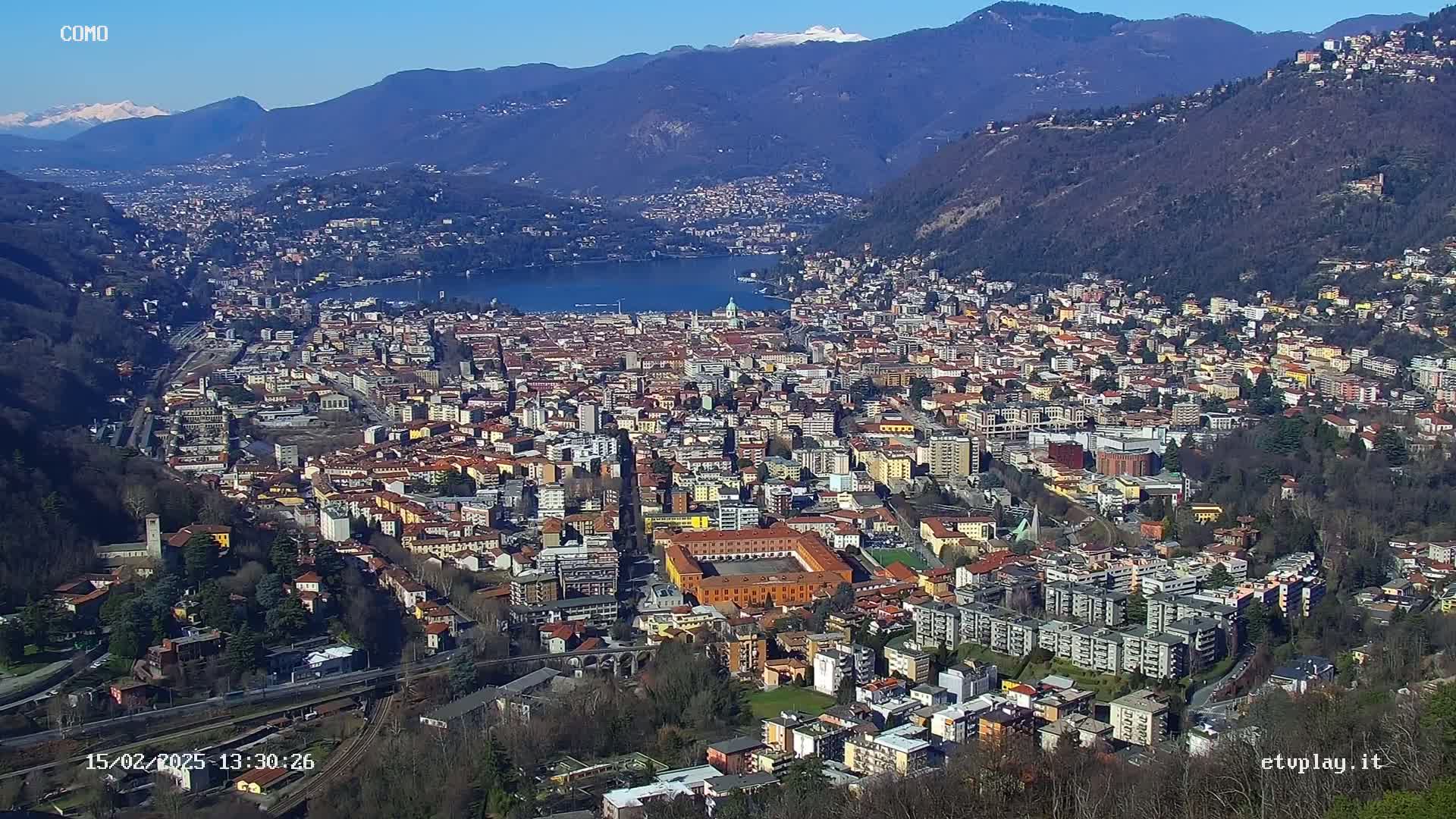 A sprawling city nestled in a valley between mountains, overlooking a lake on a sunny day with snow-capped peaks in the distance.