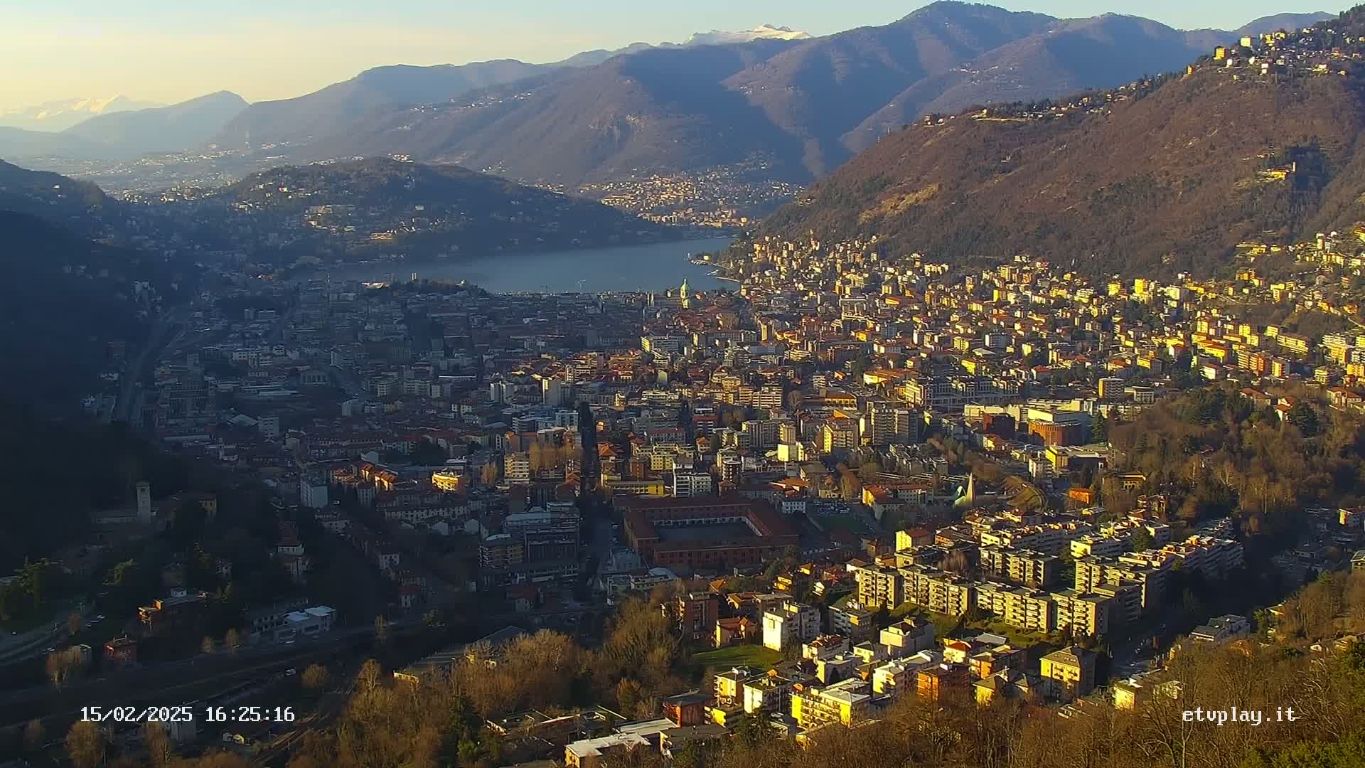A sprawling city nestled between mountains and a lake under a clear, sunny sky.