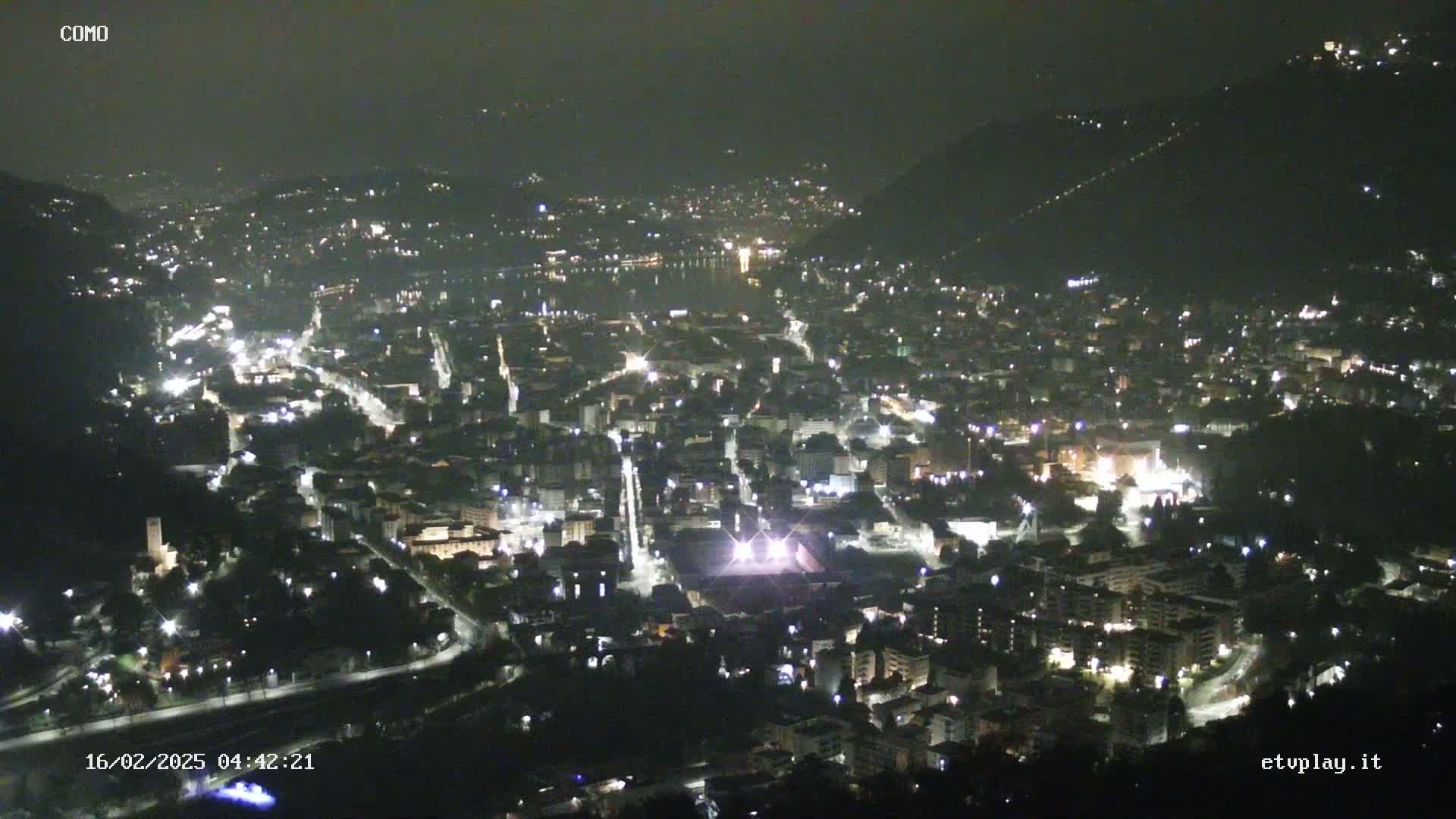 A nighttime, high-angle view of a city illuminated by streetlights, nestled between hills, under a dark sky.