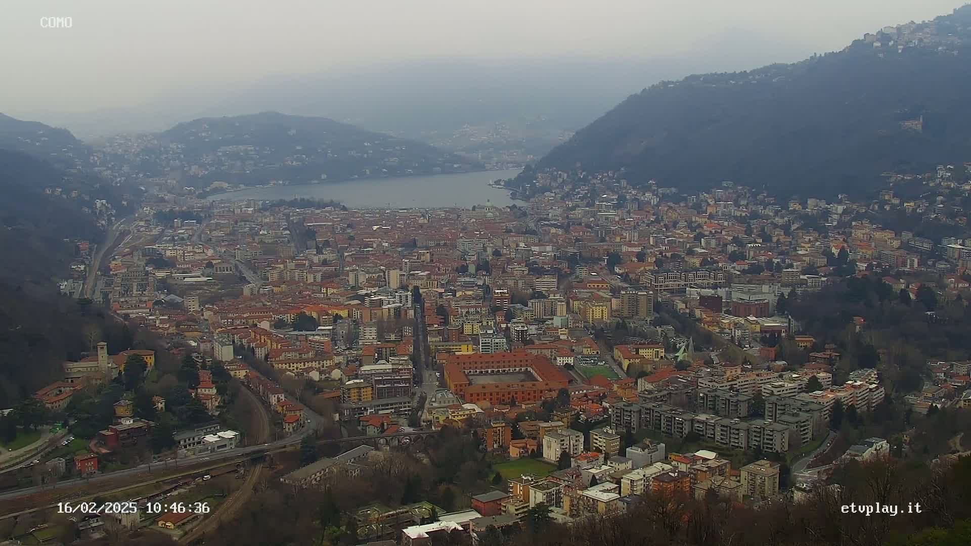 A hazy, overcast day reveals a sprawling city nestled between mountains and a lake.