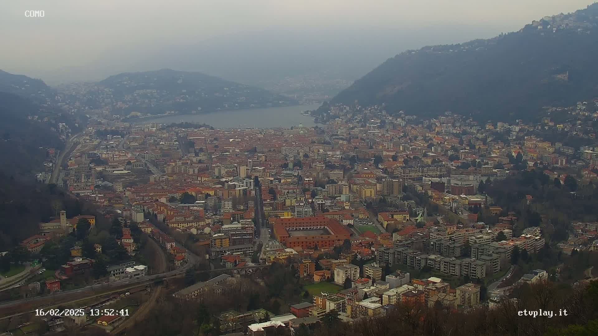 A hazy view of a large city nestled between hills and a lake, under an overcast sky.