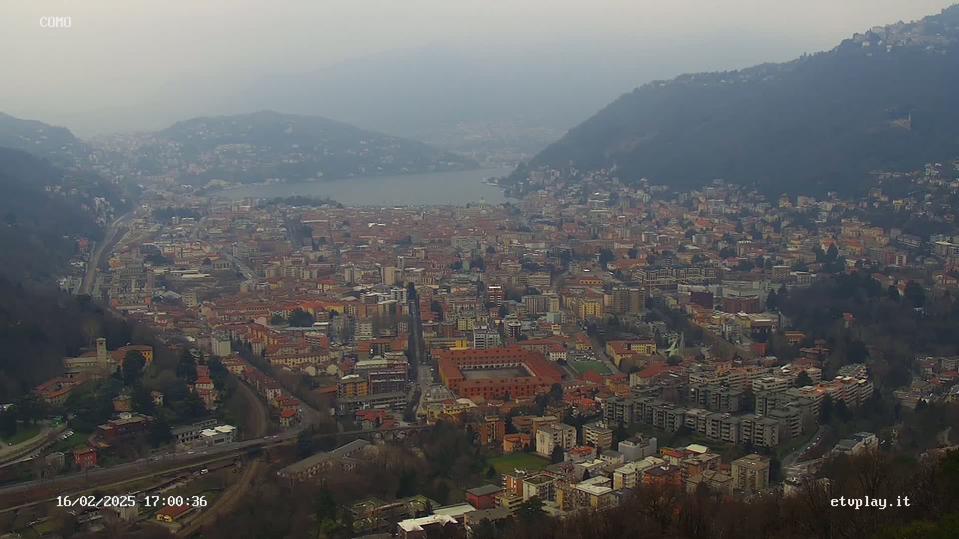A hazy, overcast view of a large city nestled between hills and a lake.